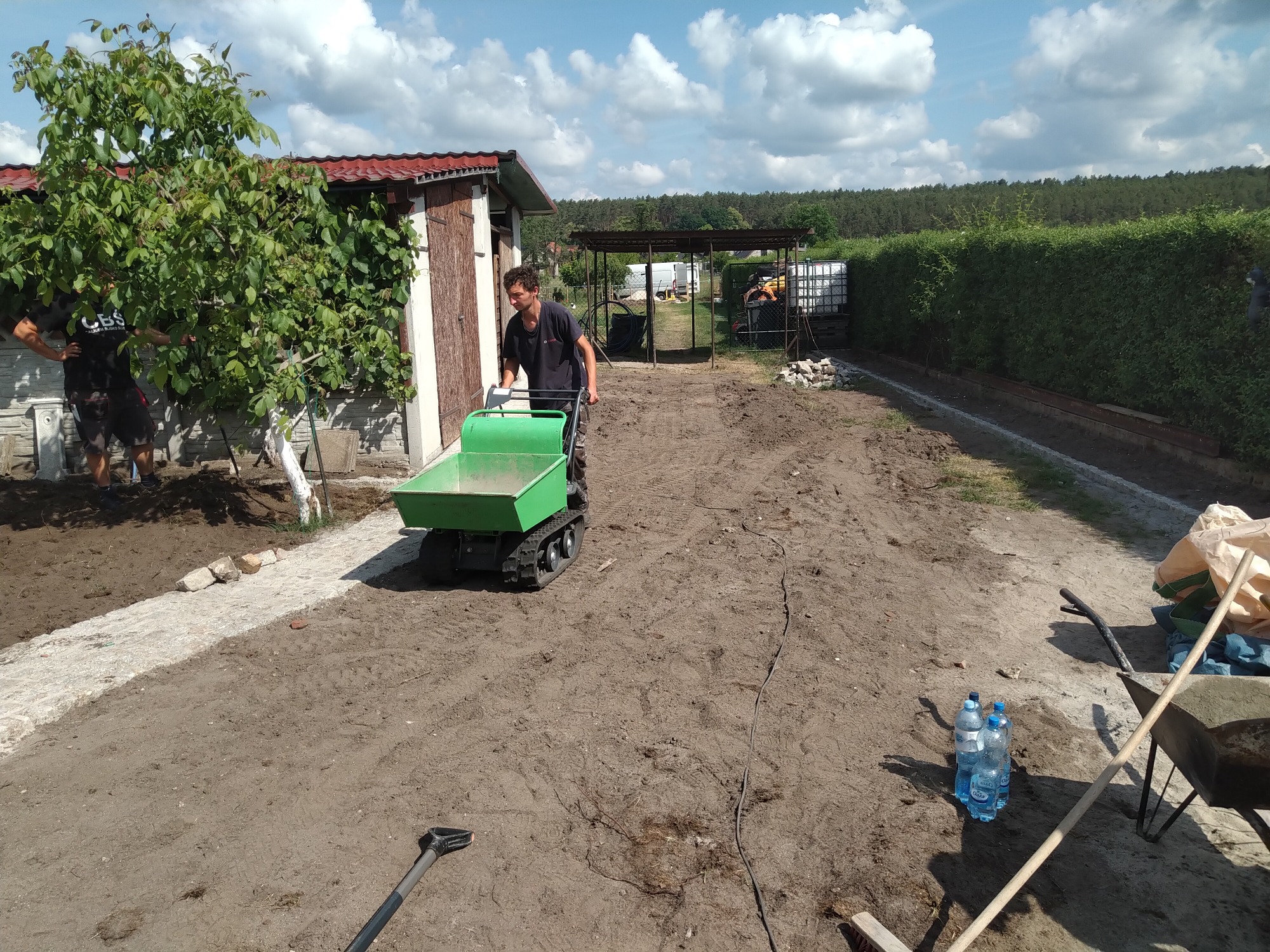 Mężczyzna prowadzi mini-wywrotkę gąsienicową po rozplantowanej ziemi na terenie posesji, widoczny budynek gospodarczy, drzewo i przygotowane obrzeże ścieżki.