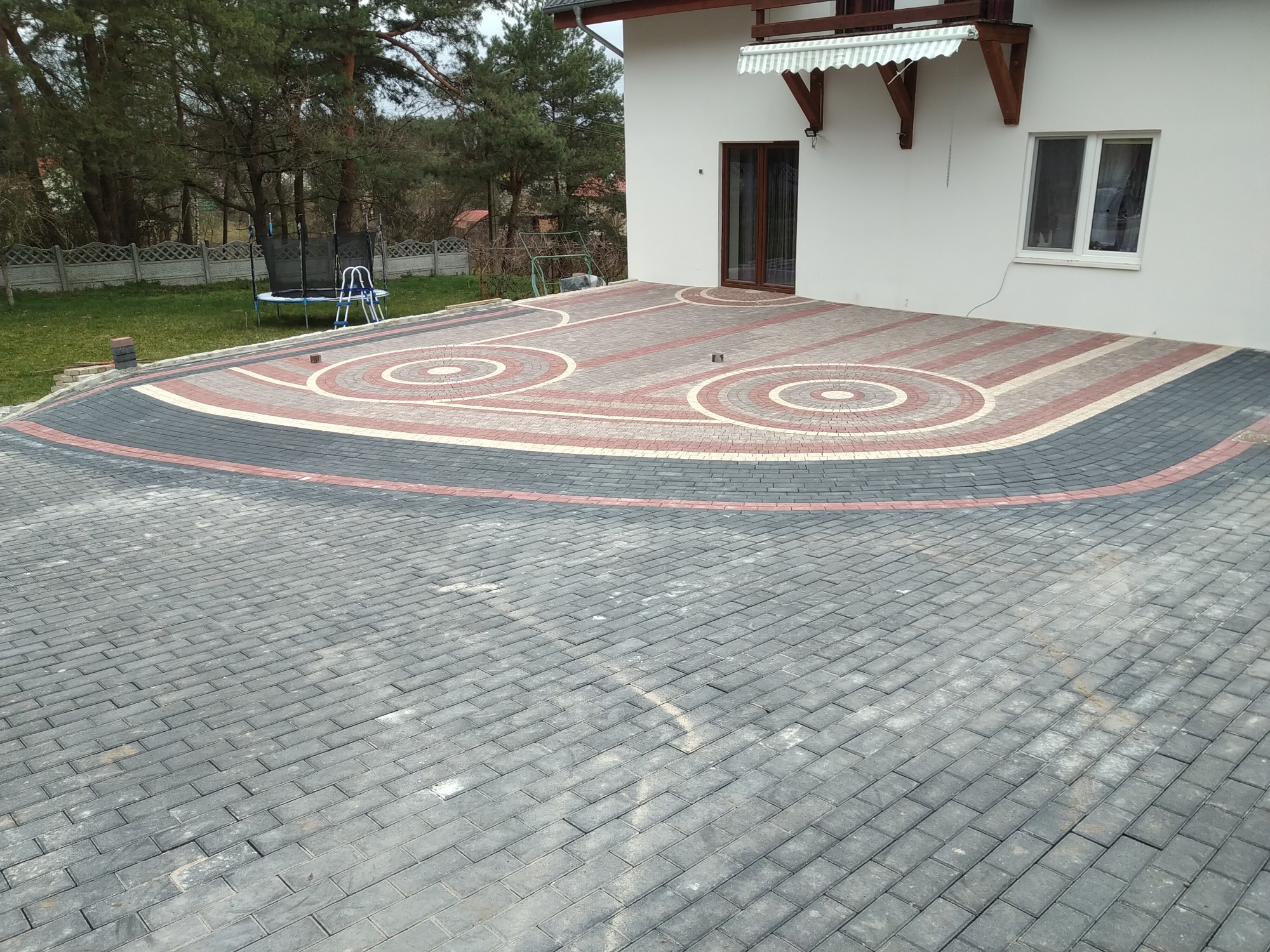 Nowo ułożona kostka brukowa w odcieniach szarości, czerwieni i bieli tworząca wzór okręgów przed białym domem z brązowymi drzwiami i oknem. W tle widoczny trawnik i trampolina.