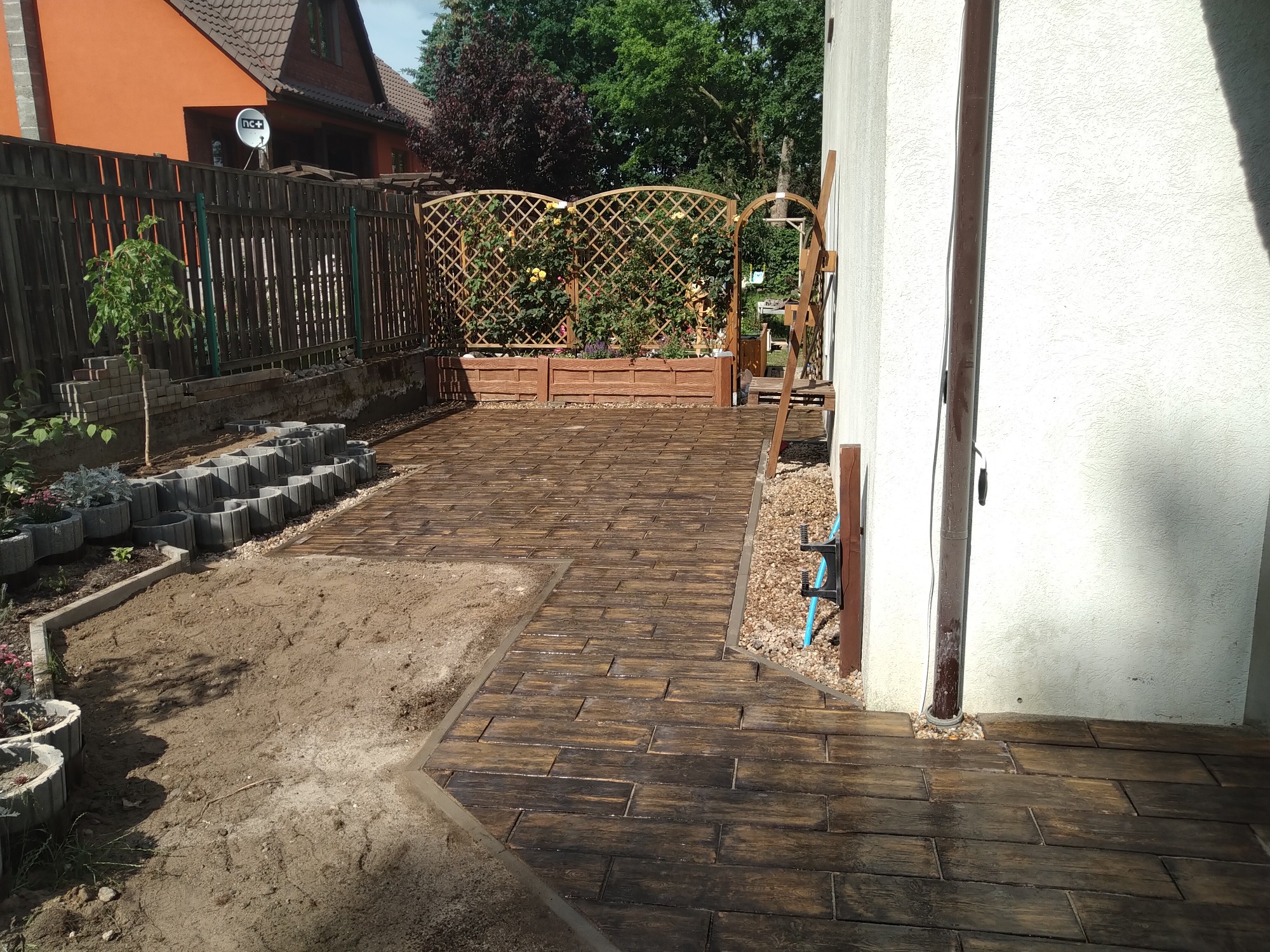 Ułożony chodnik z imitacji drewna na posesji, widoczne rabaty kwiatowe i elementy dekoracyjne ogrodu.