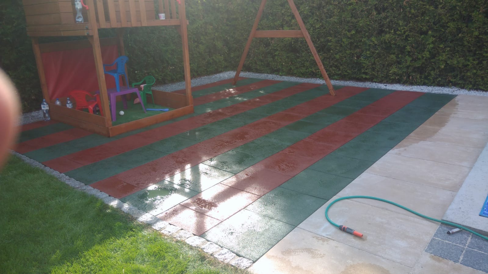 Ułożona nawierzchnia z czerwono-zielonej gumy na placu zabaw w Zielonej Górze, obok drewniana konstrukcja ze zjeżdżalnią, w tle żywopłot.