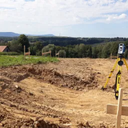 Geodezyjny tachimetr na żółtym statywie, niebieska skrzynka obok, w tle wytyczone drewniane słupki z pomarańczowymi znacznikami, dom i samochód, pagórkowaty krajobraz.