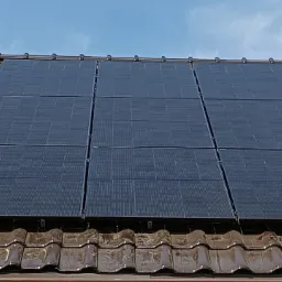 Cztery panele słoneczne zamontowane na brązowym dachu z dachówki ceramicznej, widoczne mocowania i komin w tle, błękitne niebo.