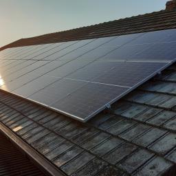 EL-MAR Instalacje elektryczne i fotowoltaiczne Mariusz Kordek - Panele słoneczne zamontowane na dachu z dachówki ceramicznej, widoczne mocowania, w tle zachodzące słońce i sylwetka osoby na drabinie.