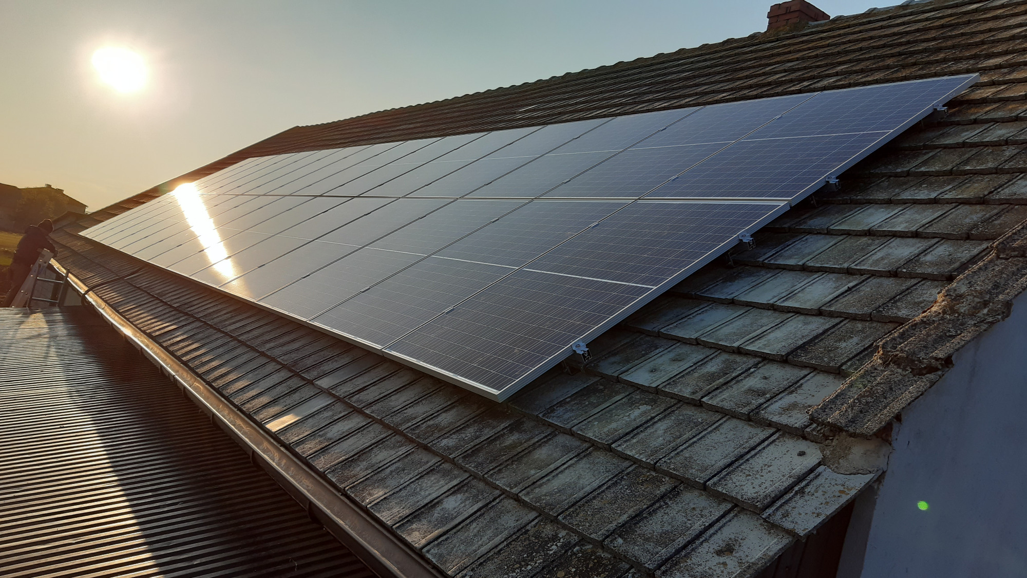 Panele słoneczne zamontowane na dachu z dachówki ceramicznej, widoczne mocowania, w tle zachodzące słońce i sylwetka osoby na drabinie.