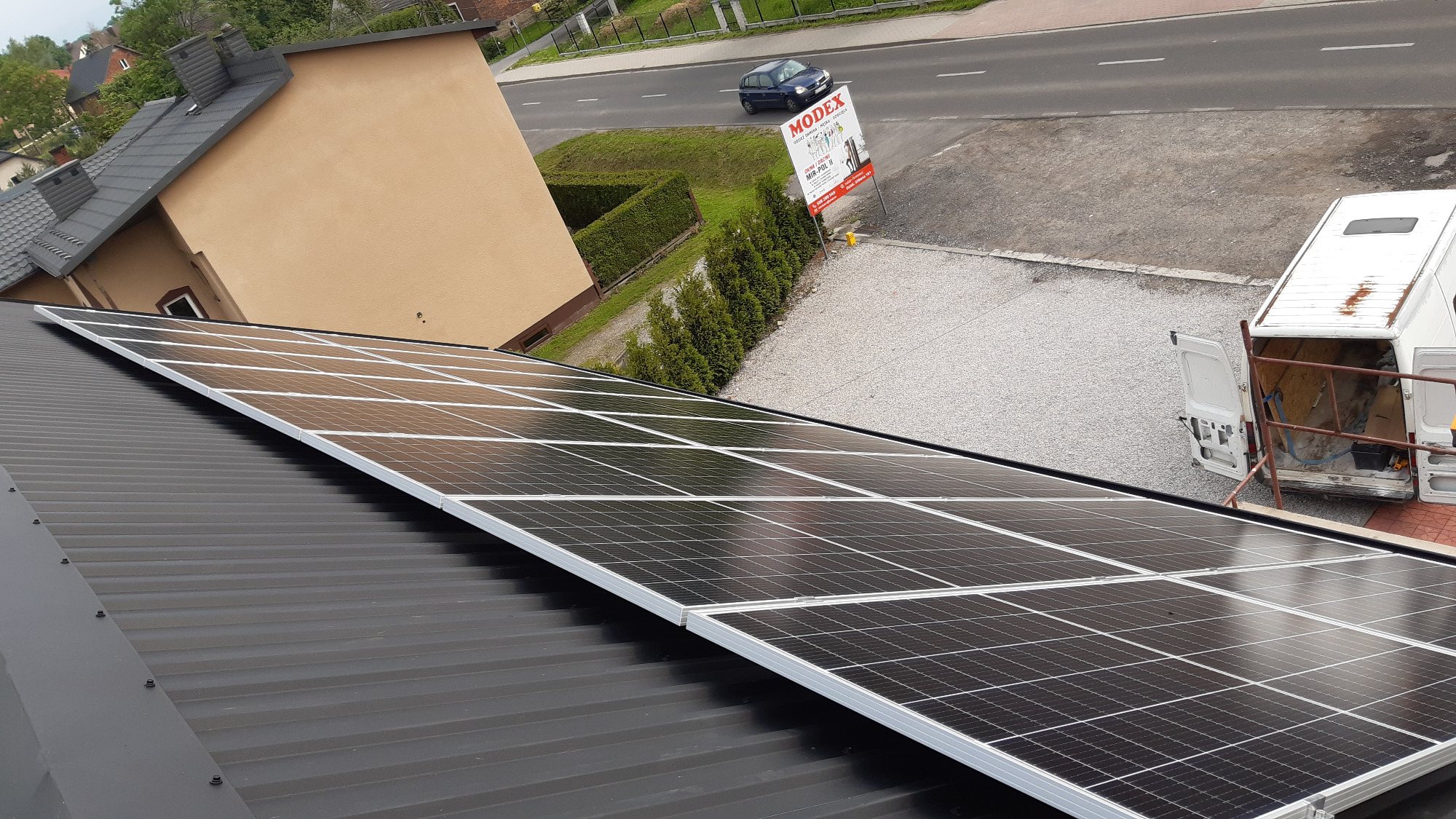 Panel słoneczny na czarnym dachu blaszanej konstrukcji, widok z góry na samochód dostawczy z otwartymi drzwiami w tle.