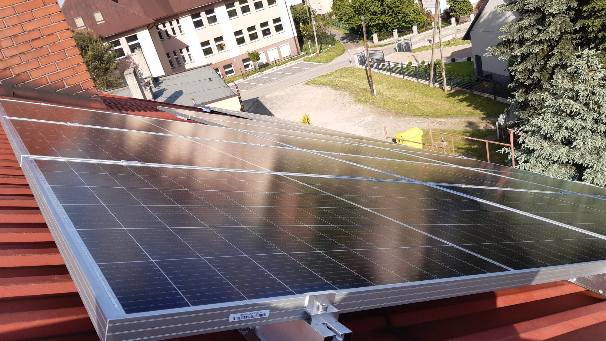 Panele słoneczne na dachu z czerwonej dachówki, widok z góry na budynek szkolny i otoczenie.
