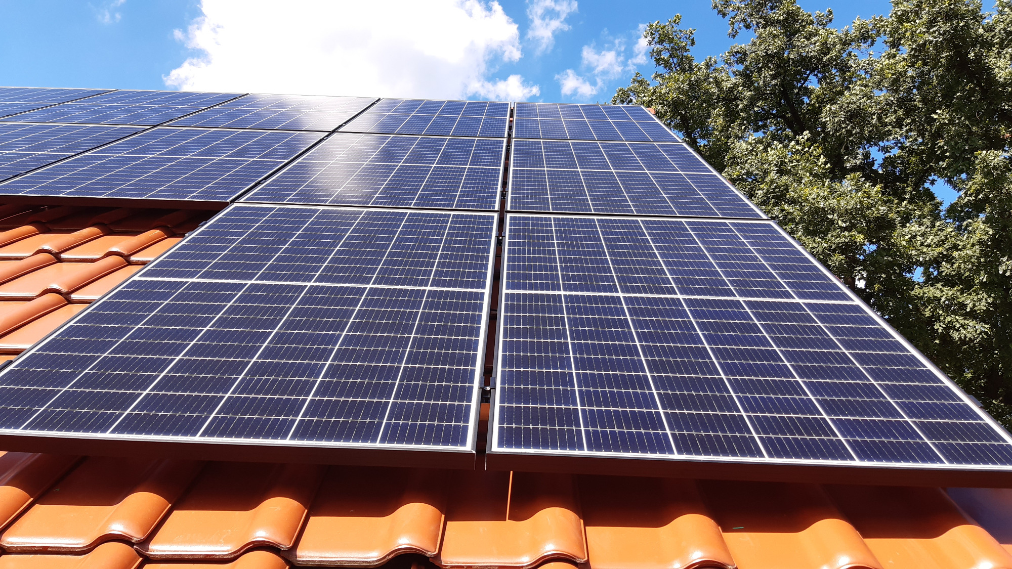 Panele słoneczne na dachu z czerwonej dachówki, widoczne na tle błękitnego nieba z chmurami i zieleni drzew.