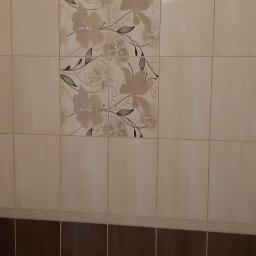 Lilbud Project - Beżowe i brązowe płytki ścienne w łazience, z dekoracyjnym motywem kwiatowym na części płytek, oraz widoczne zawory na dole ściany.