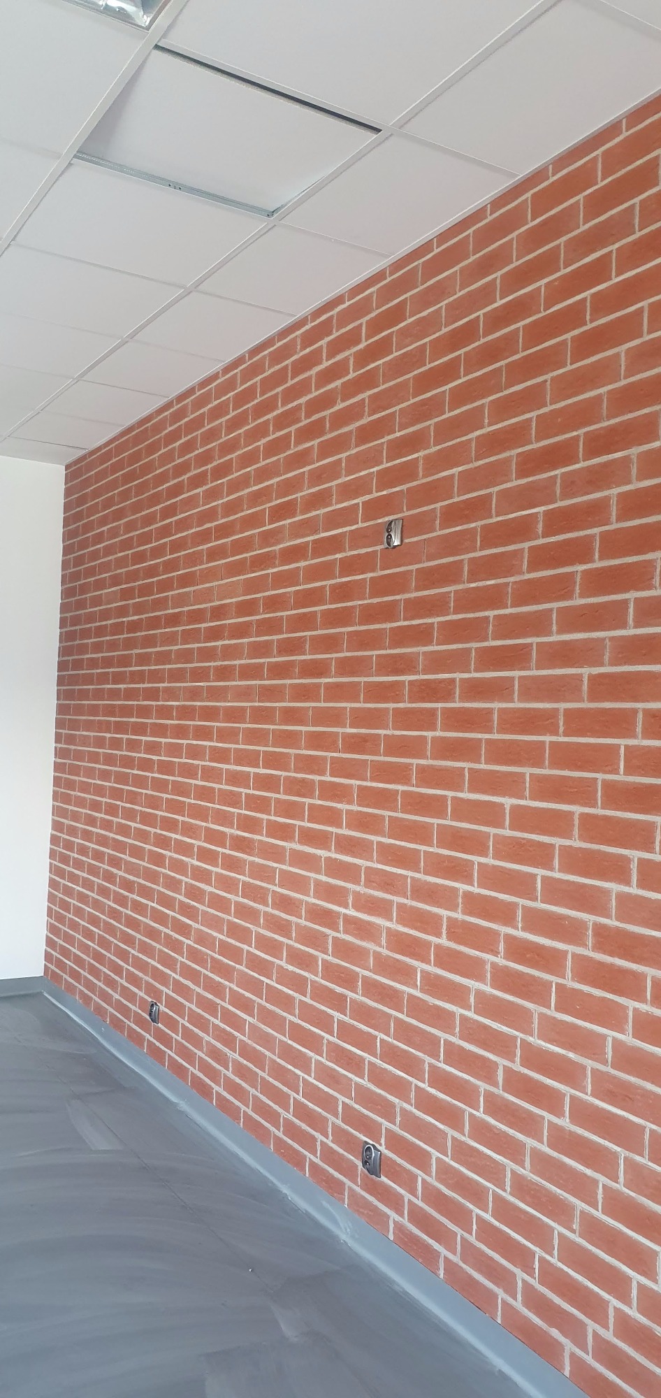 Ściana z cegły klinkierowej w pomieszczeniu biurowym, widoczne gniazdka elektryczne, szara podłoga i sufit podwieszany.