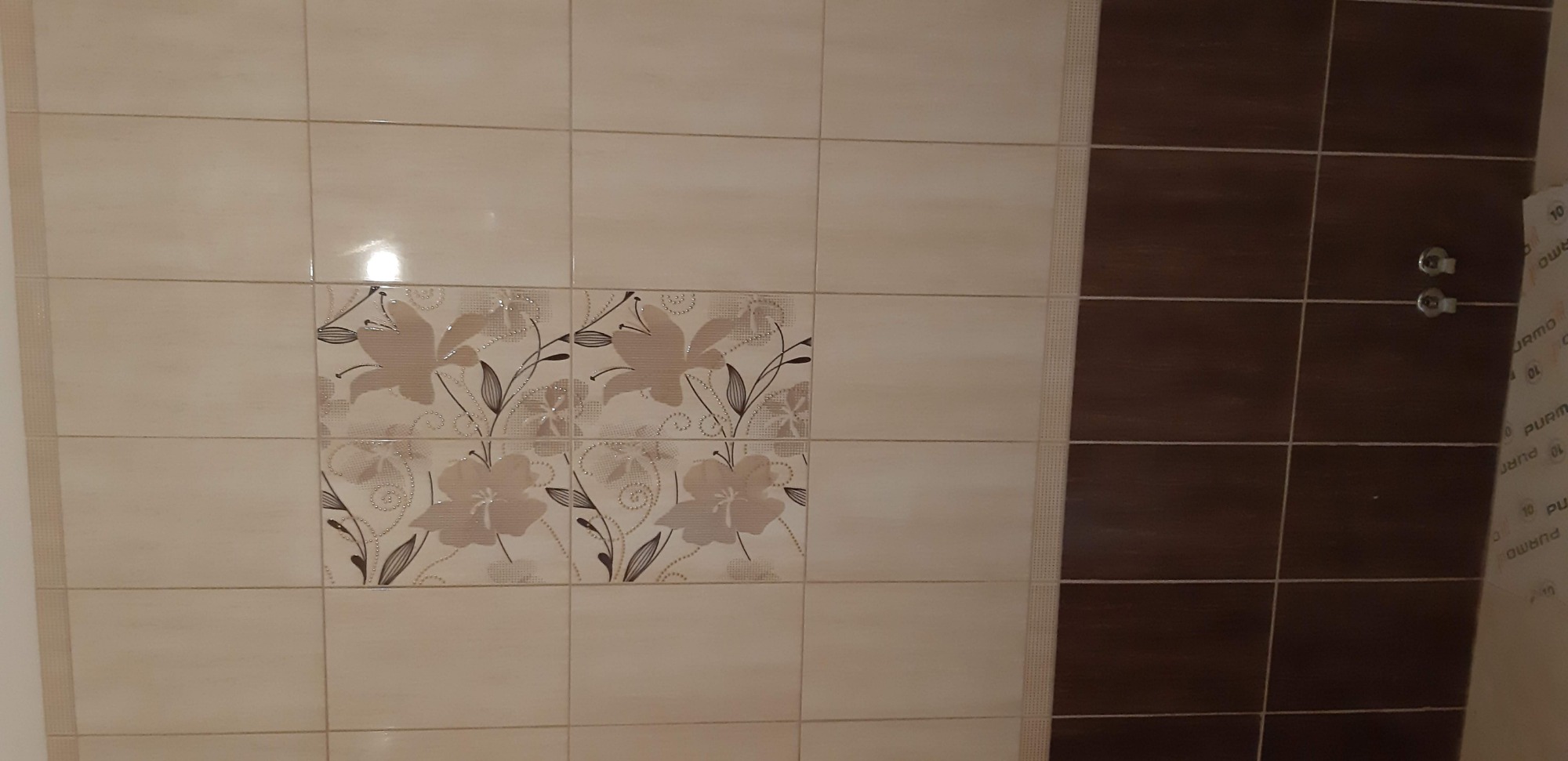 Ściana wyłożona beżowymi i brązowymi płytkami ceramicznymi z motywem kwiatowym, widoczne dwa chromowane zawory.