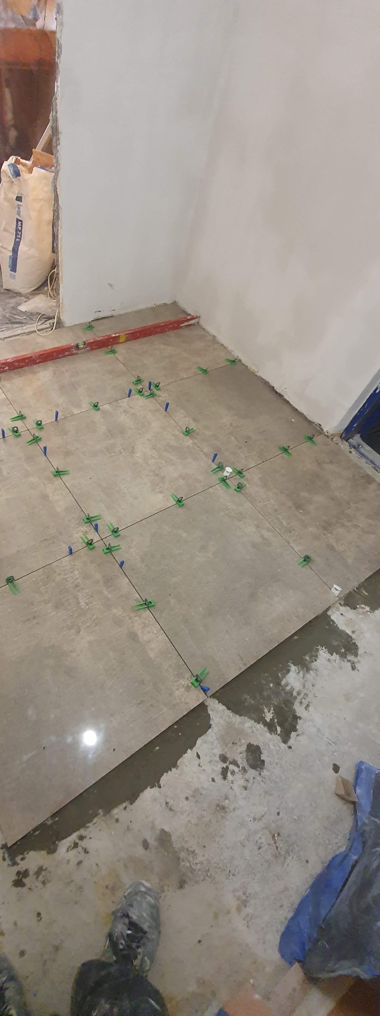 Podłoga w trakcie układania płytek, widoczne krzyżyki dystansowe i poziomica, worek zaprawy w tle, fragment obuwia robotnika w dolnej części kadru.