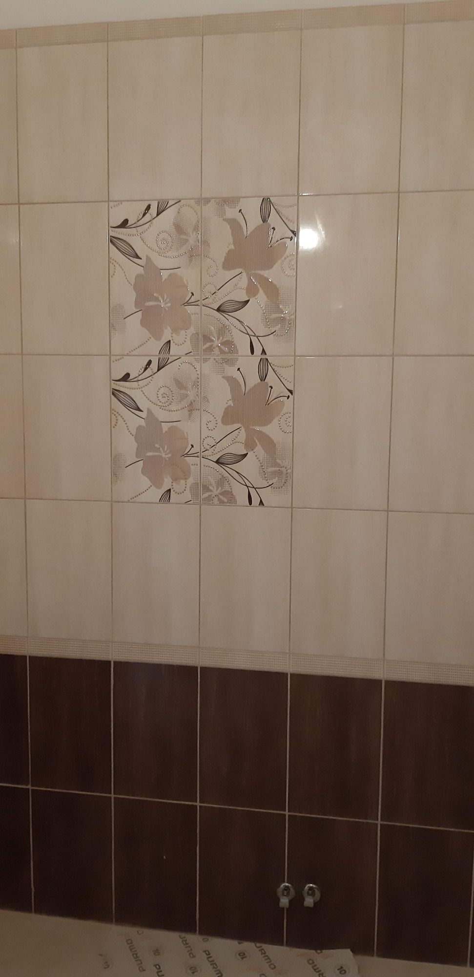 Beżowe i brązowe płytki ścienne w łazience, z dekoracyjnym motywem kwiatowym na części płytek, oraz widoczne zawory na dole ściany.