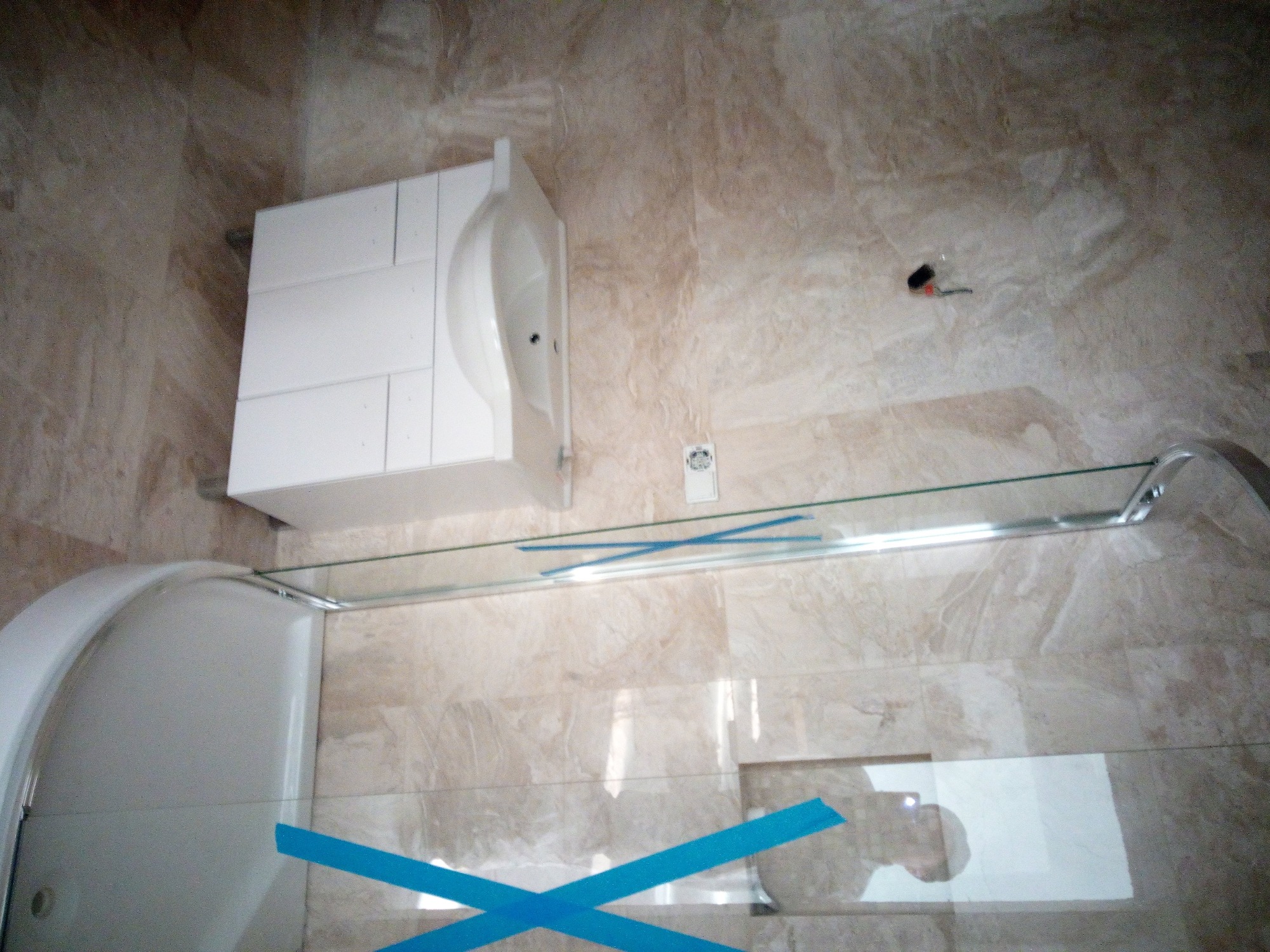 Wykończona łazienka z beżowymi płytkami imitującymi marmur, białą szafką z umywalką, szklaną kabiną prysznicową zabezpieczoną niebieską taśmą oraz szklaną półką.