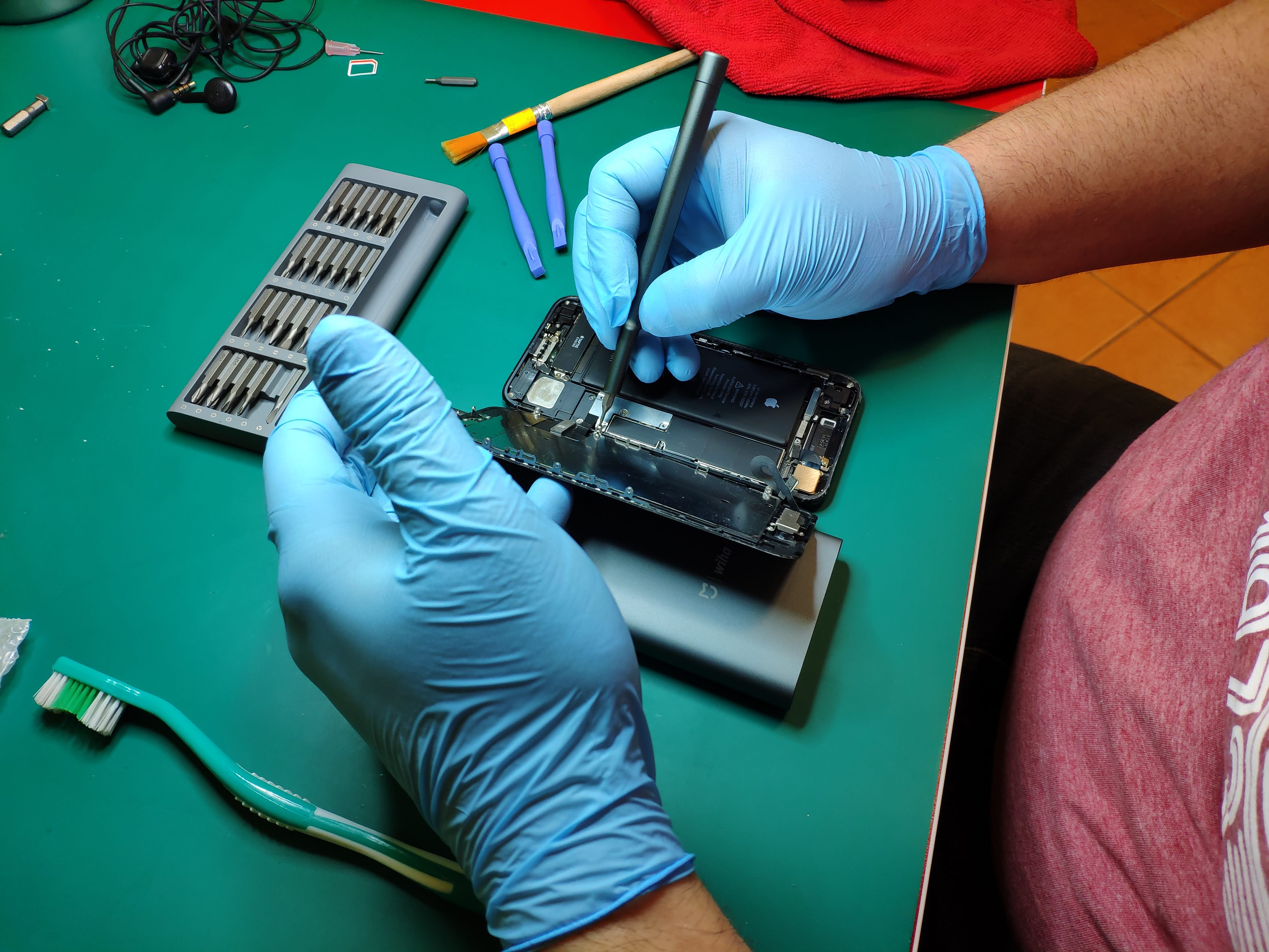 Otwarty smartfon z widoczną baterią, naprawiany przy użyciu precyzyjnego śrubokręta w rękawicach ochronnych na zielonym blacie, obok zestaw narzędzi, pędzel i szczoteczka do czyszczenia.