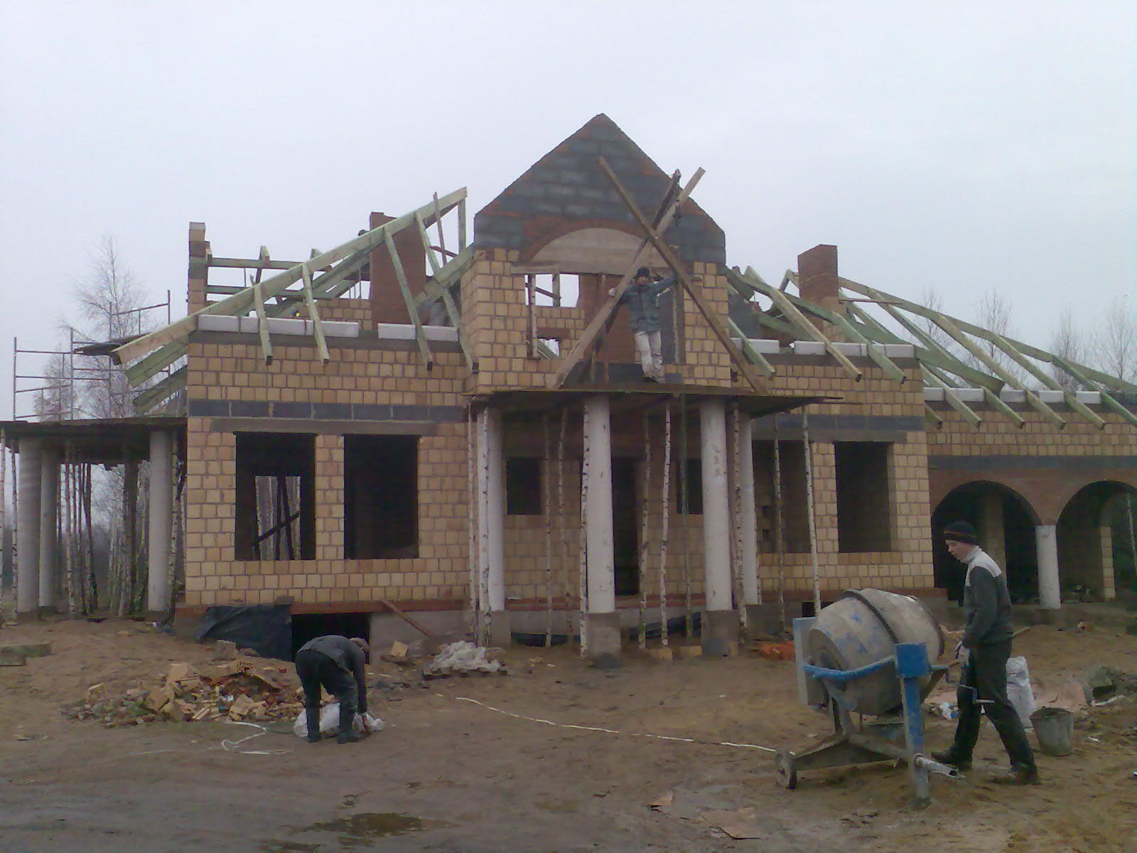 Budowa domu w stanie deweloperskim: surowa konstrukcja z cegły, widoczne elementy dachu z drewnianych belek, pracownik na rusztowaniu i betoniarka na pierwszym planie, pochmurne niebo w tle.