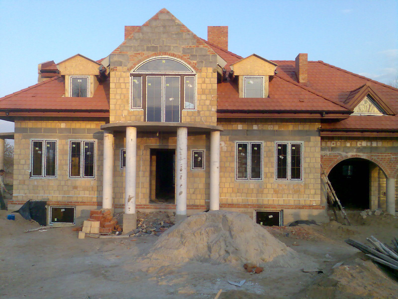 Budynek jednorodzinny w stanie deweloperskim: surowa elewacja z żółtej cegły, czerwona dachówka ceramiczna, widoczne okna z białymi ramami oraz kolumny podtrzymujące balkon, na pierwszym planie...