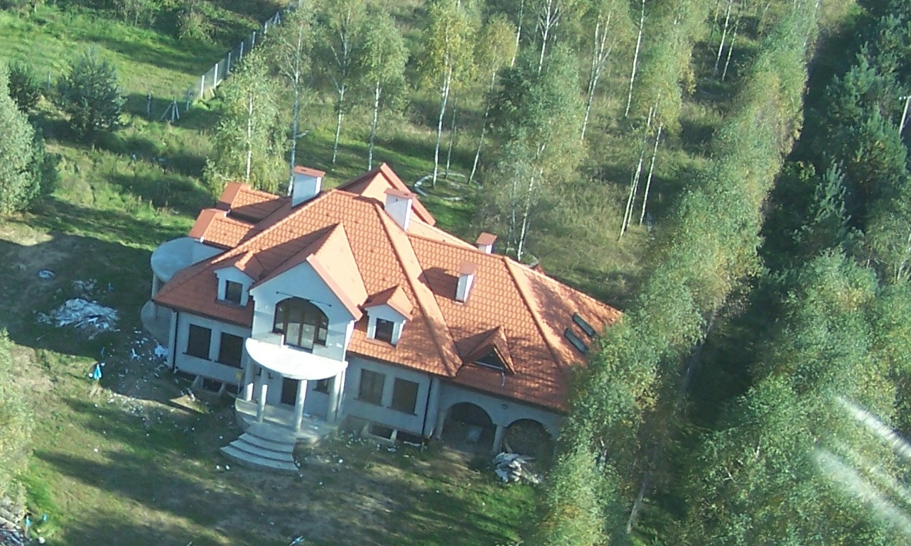 Dom jednorodzinny w stanie deweloperskim z pomarańczową dachówką, widok z góry, otoczony drzewami, z widocznymi śladami budowy na trawniku.