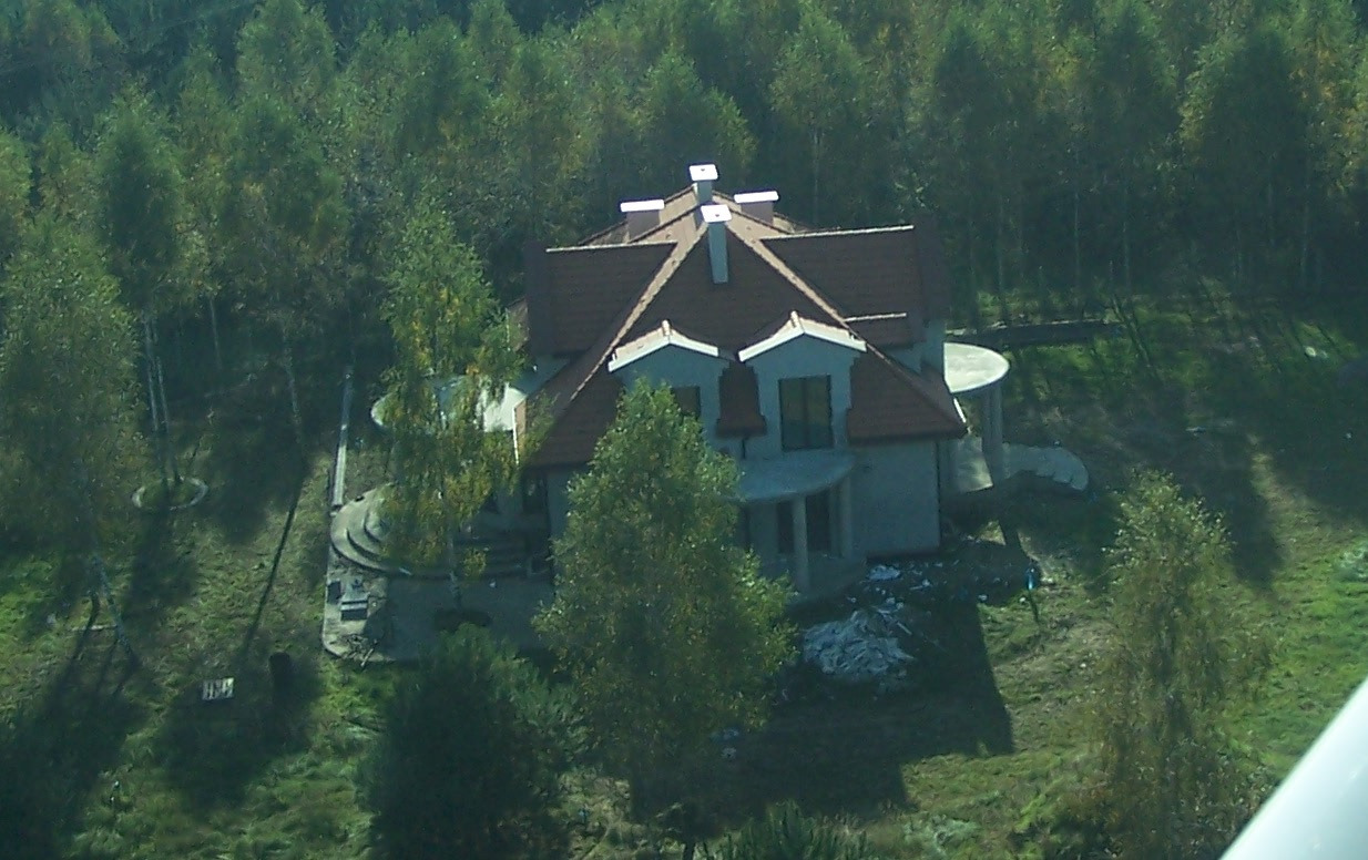 Widok z góry na budynek w stanie deweloperskim z ciemnoczerwonym dachem, otoczony zielenią drzew i trawników. Elementy elewacji w kolorze szarym, widoczne rusztowania i materiały budowlane.