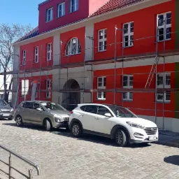 Budynek w trakcie renowacji z rusztowaniami, pomalowany na czerwono i biało, z zaparkowanymi samochodami na brukowanej ulicy.