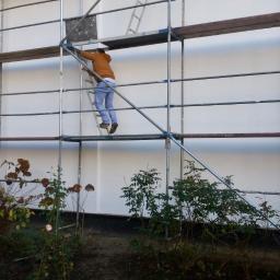 Molbud - Pracownik w brązowej kurtce wchodzi po drabince na rusztowaniu ustawionym przy białej ścianie budynku, widoczne rośliny w ogrodzie u dołu.