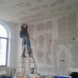 Molbud - Pracownik na drabinie szpachluje łączenia płyt kartonowo-gipsowych na suficie i ścianach w trakcie prac wykończeniowych w remontowanym pomieszczeniu, widoczne narzędzia i materiały budowlane.