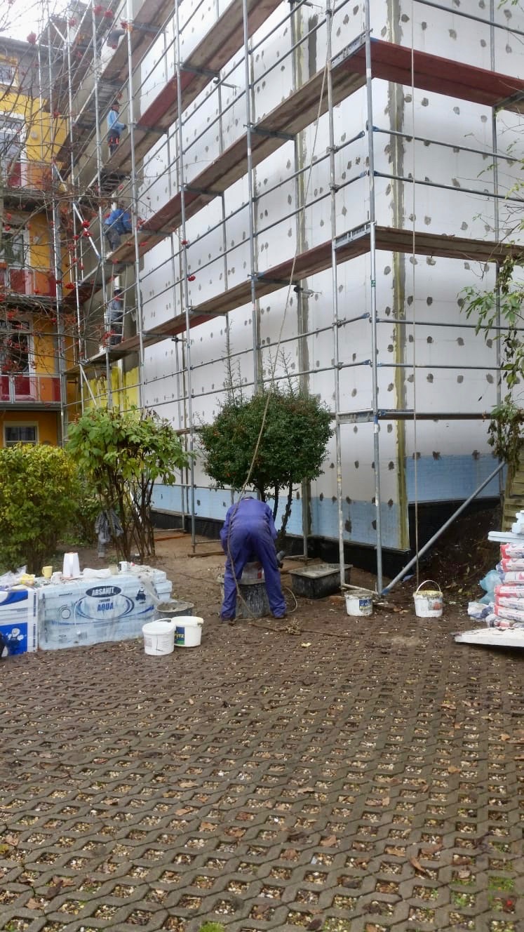 Pracownik w niebieskim kombinezonie miesza zaprawę na budowie, widoczne rusztowania i ocieplone ściany budynku z białym styropianem, opakowania materiałów budowlanych i drzewa w tle.