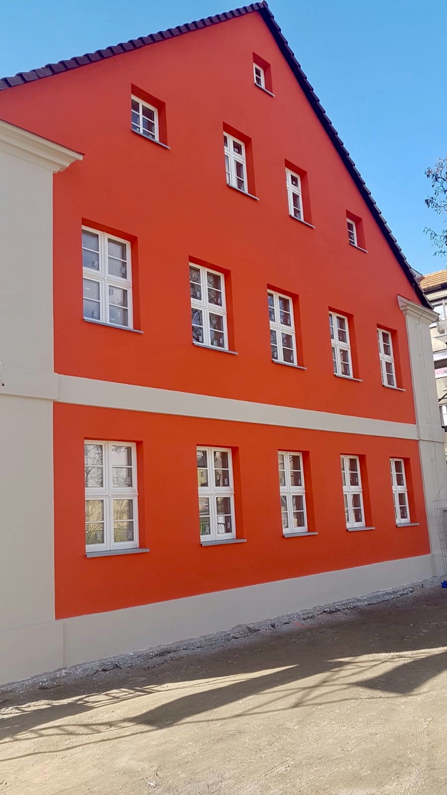 Odnowiona fasada budynku z pomarańczową ścianą i białymi oknami, widoczne elementy wykończeniowe w kolorze kremowym.