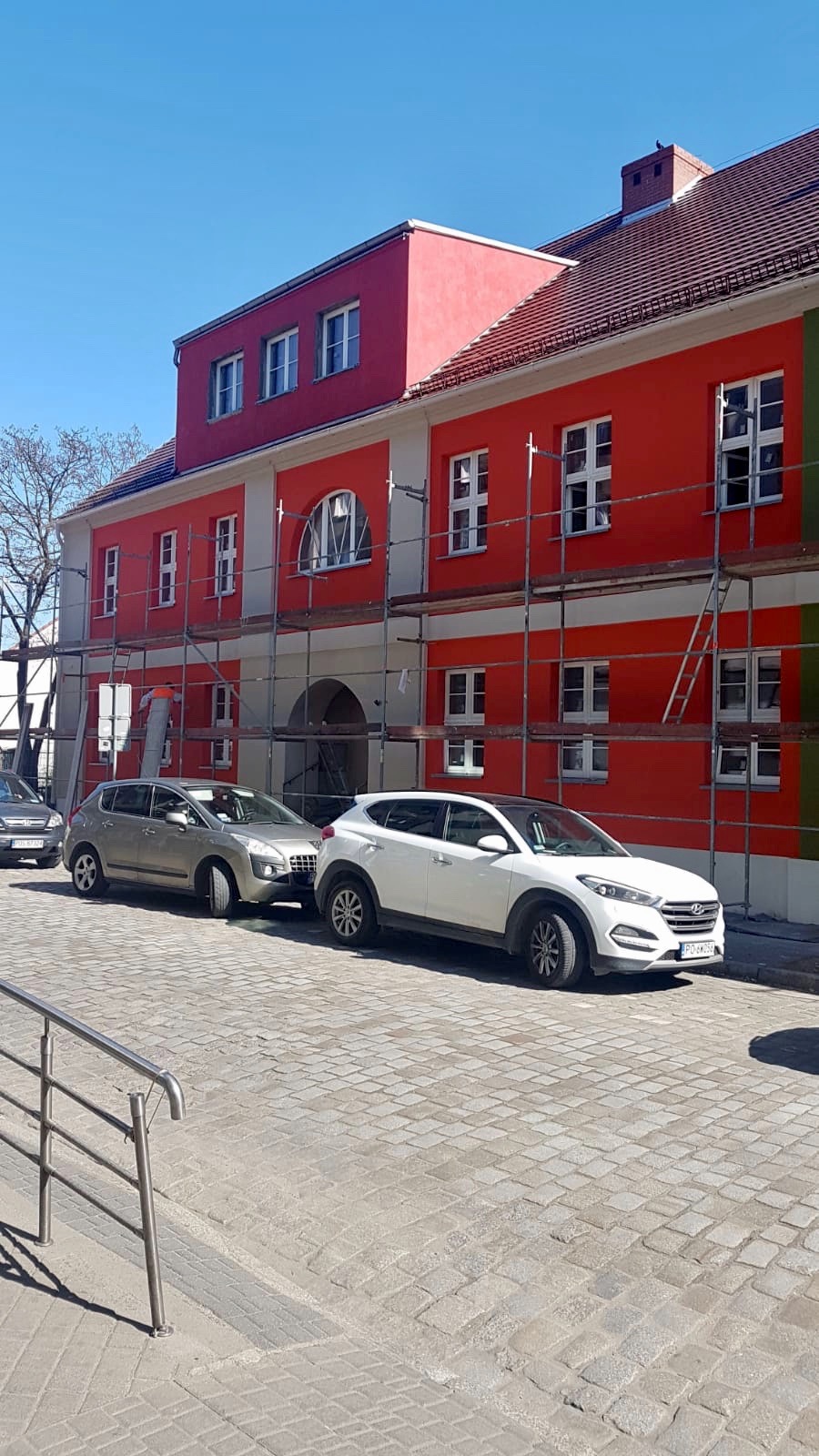 Budynek w trakcie renowacji z rusztowaniami, pomalowany na czerwono i biało, z zaparkowanymi samochodami na brukowanej ulicy.