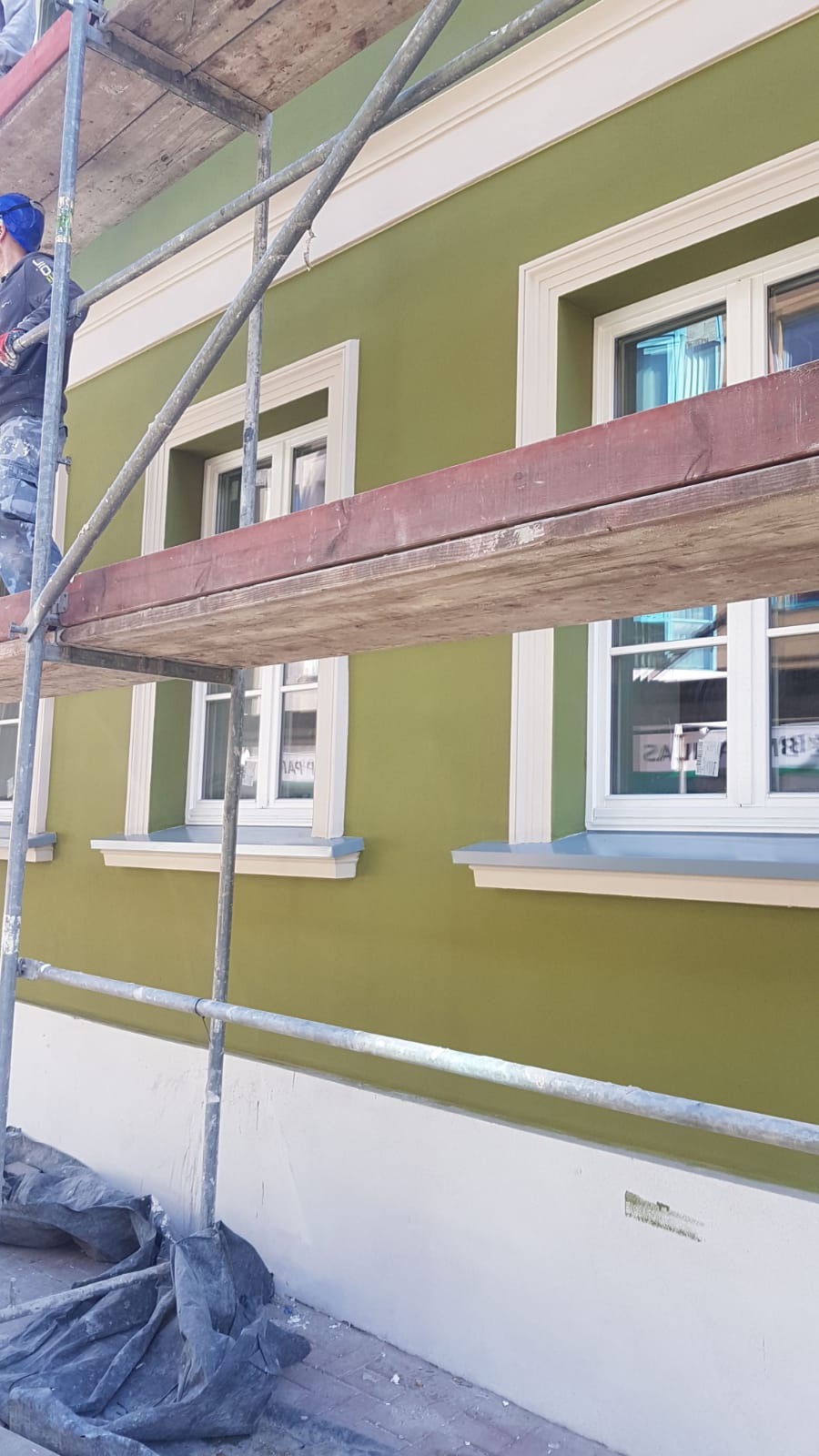 Pracownik na rusztowaniu maluje elewację budynku na oliwkowy kolor, widoczne białe obramowania okien i dolna część ściany w jasnym odcieniu.