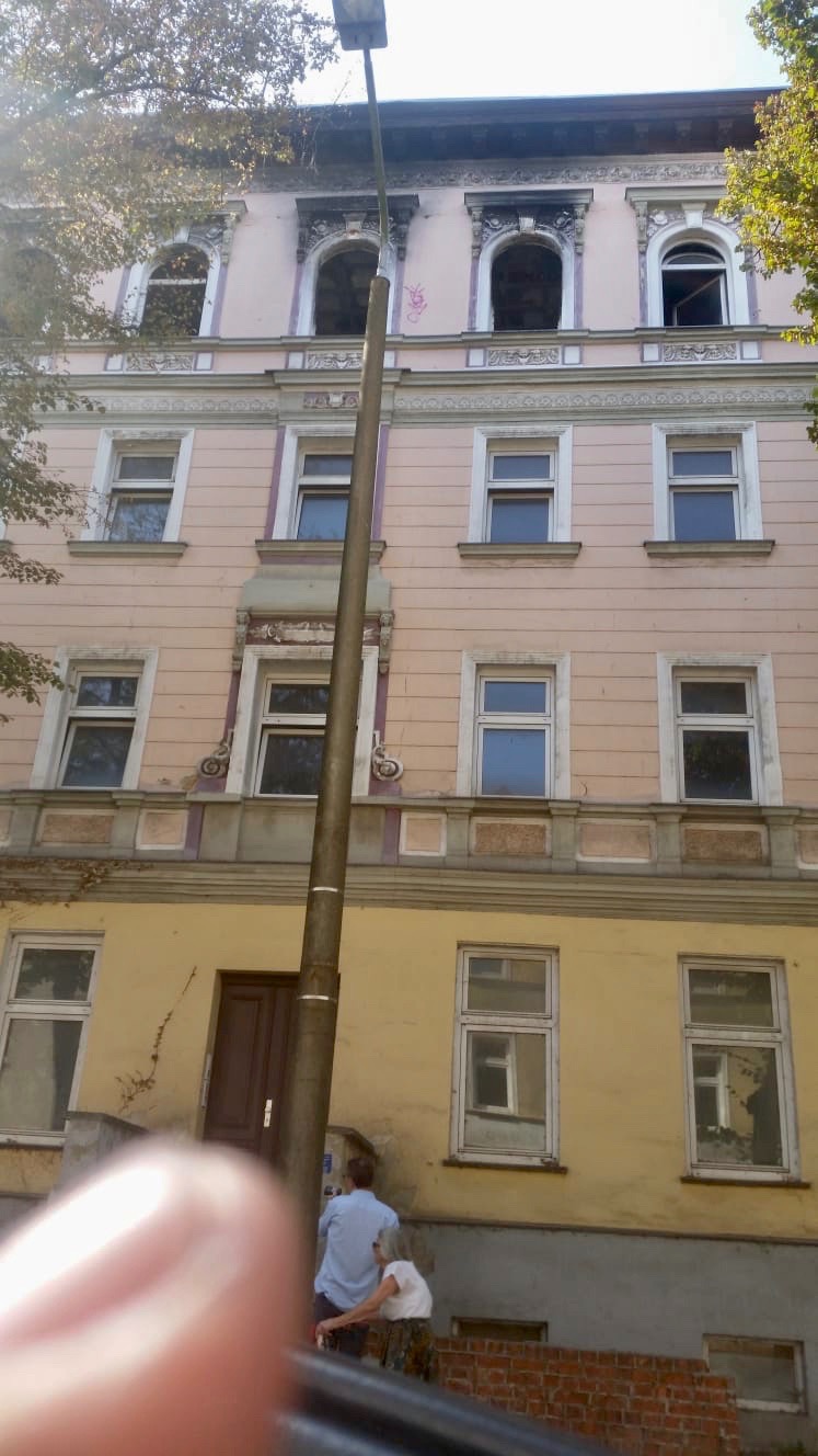 Elewacja kamienicy przed remontem, widoczne okna z drewnianą stolarką, ślady po pożarze na górnych oknach, stara lampa uliczna i dwie osoby na pierwszym planie