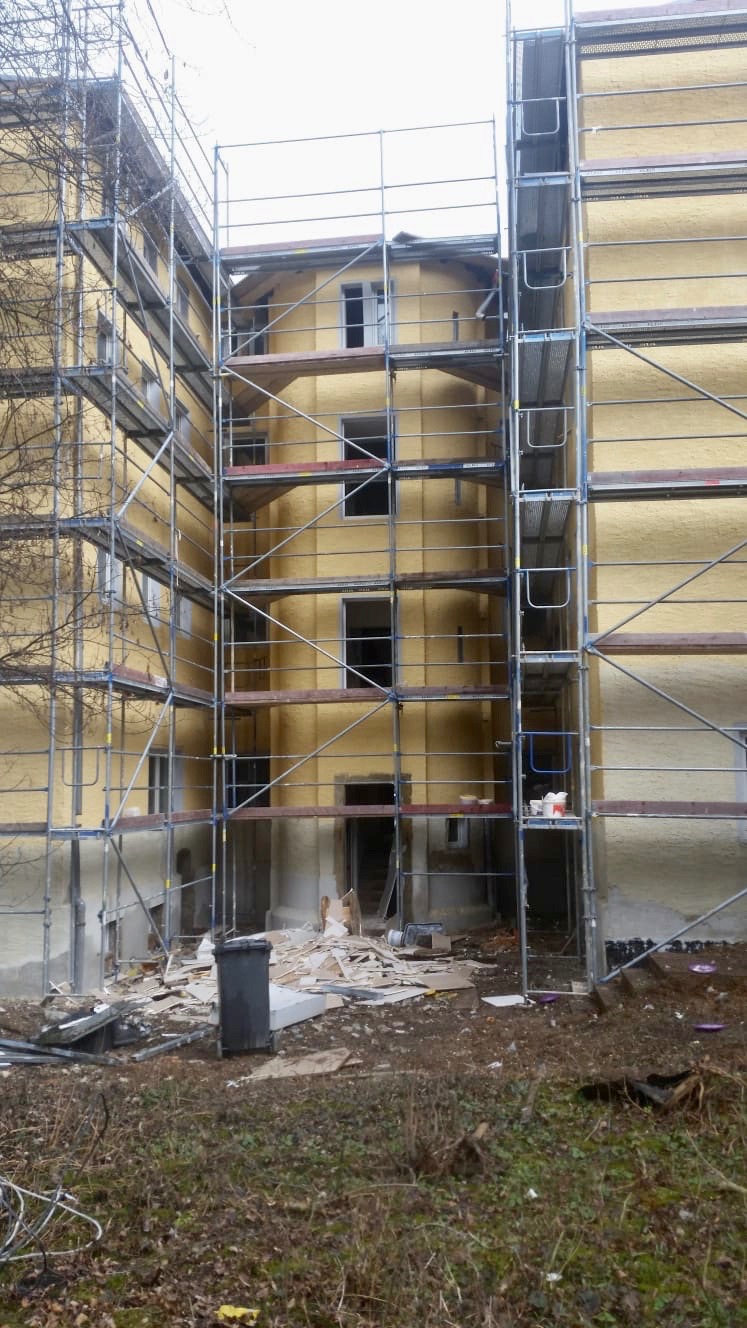 Budynek w trakcie remontu, otoczony rusztowaniami, widoczne ślady prac i gruz.