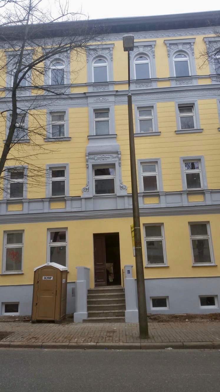 Żółta kamienica z szarymi elementami dekoracyjnymi, widoczna przenośna toaleta przed wejściem, wskazująca na trwający remont elewacji.