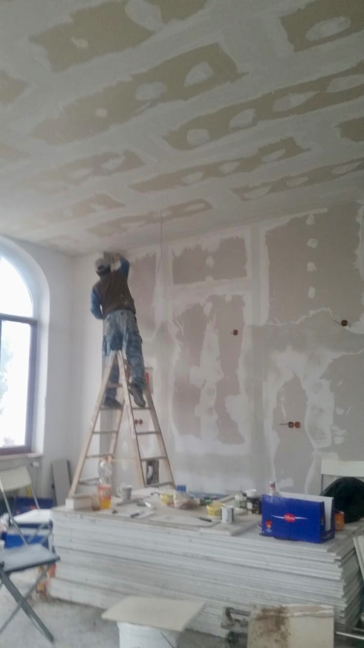 Pracownik na drabinie szpachluje łączenia płyt kartonowo-gipsowych na suficie i ścianach w trakcie prac wykończeniowych w remontowanym pomieszczeniu, widoczne narzędzia i materiały budowlane.