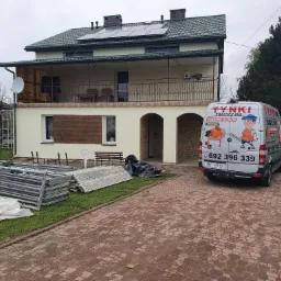 Dwukondygnacyjny dom z ocieploną elewacją, widoczne rusztowania i samochód firmy tynkarskiej z reklamą.