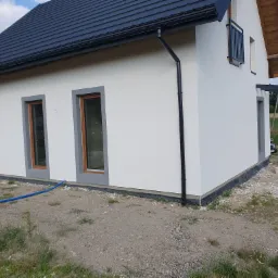 Fragment elewacji domu jednorodzinnego z widoczną strukturą tynku, grafitowymi obramowaniami okien i czarną rynną spustową.