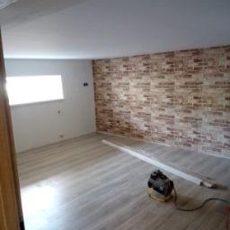 k&m rem - Pokój po remoncie: ściana z cegły dekoracyjnej, jasna podłoga z paneli, świeżo pomalowany sufit, widoczny grzejnik elektryczny i listwy przypodłogowe do montażu.