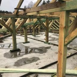 k&m rem - Wznoszenie drewnianej konstrukcji dachu na betonowej płycie fundamentowej, widoczne drewniane słupy i krokwie, mokra powierzchnia betonu.