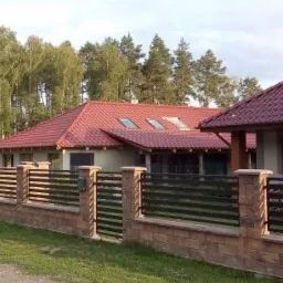 Dom jednorodzinny z czerwonym dachem i oknami dachowymi, ogrodzony płotem z ceglanymi słupkami i metalowymi przęsłami, otoczony zielenią.