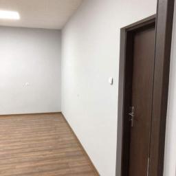 k&m rem - Pomalowane na biało wnętrze z drewnopodobną podłogą, brązowymi listwami przypodłogowymi i ciemnymi drewnianymi drzwiami z srebrną klamką po prawej stronie.