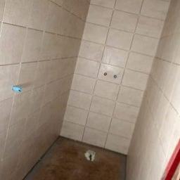 k&m rem - Wnętrze łazienki w trakcie prac: ściany wyłożone jasnymi, kwadratowymi płytkami, widoczne przyłącza wodne, podłoga wylewka, fragment drabiny malarskiej w rogu.