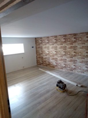 Pokój po remoncie: ściana z cegły dekoracyjnej, jasna podłoga z paneli, świeżo pomalowany sufit, widoczny grzejnik elektryczny i listwy przypodłogowe do montażu.