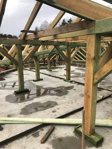 Wznoszenie drewnianej konstrukcji dachu na betonowej płycie fundamentowej, widoczne drewniane słupy i krokwie, mokra powierzchnia betonu.