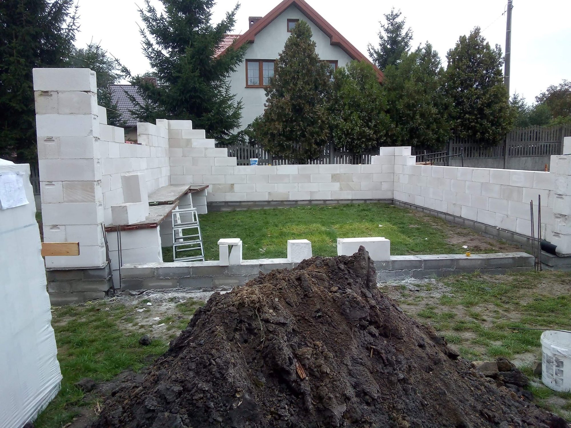 Wznoszenie ścian fundamentowych z bloczków betonowych, widoczny stos ziemi, drabina i częściowo ukończona konstrukcja z widokiem na dom z czerwonym dachem w tle.