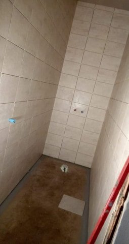Wnętrze łazienki w trakcie prac: ściany wyłożone jasnymi, kwadratowymi płytkami, widoczne przyłącza wodne, podłoga wylewka, fragment drabiny malarskiej w rogu.