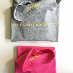 Dwie złożone koszulki leżące jedna na drugiej: szara i różowa, obie z żółtym napisem 'Taka koszulkę mam tylko ja' oraz logo marki na rękawie.