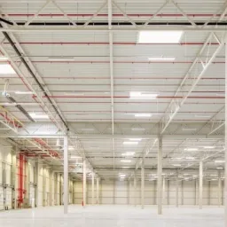 Wnętrze pustej hali magazynowej z betonowymi filarami, systemem rur pod sufitem i bramami rolowanymi po lewej stronie.