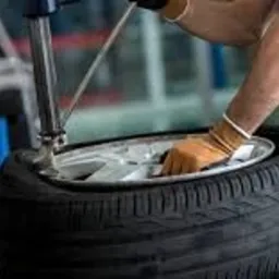 Mechanik w niebieskim kombinezonie wymienia oponę na maszynie w warsztacie, widoczny fragment felgi i rękawica ochronna.