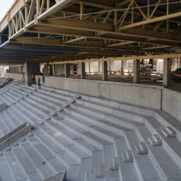 Adirect Adam Krzywdziński - Betonowe trybuny w trakcie budowy stadionu, widoczne elementy konstrukcji stalowej dachu i pracujący robotnicy w oddali.