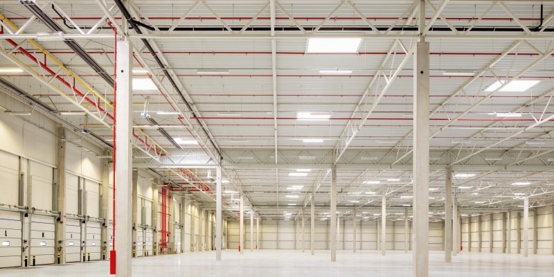 Wnętrze pustej hali magazynowej z betonowymi filarami, systemem rur pod sufitem i bramami rolowanymi po lewej stronie.