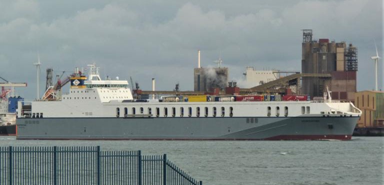 Duży statek towarowy typu Ro-Ro płynący po wodzie, z widocznymi kontenerami na pokładzie, w tle zabudowania portowe i turbina wiatrowa.